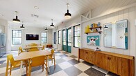 Located in Clermont, Florida, this inviting community dining and kitchen space combines modern farmhouse aesthetics with a playful, contemporary flair. The open single-floor layout accommodates multiple dining tables paired with cheerful yellow chairs, creating a welcoming communal atmosphere throughout.

The space is defined by its striking black-and-white checkered terrazzo flooring, which anchors the room with a bold, retro-inspired character. White shiplap walls and a matching paneled ceiling lend a clean, cottage-like charm, while structural white beams add architectural interest overhead. Pendant lighting with matte black fixtures provides focused illumination across the dining area.

Natural light flows generously through multiple black-framed windows, complemented by teal-painted French doors that open to outdoor views, creating a bright and airy environment. A wall-mounted TV and a chalkboard panel offer functional touches for events or gatherings, while open wooden shelves and a warm wood credenza add rustic character to the dining side of the room.

The fully equipped kitchen features crisp white shaker cabinetry, dark granite-style countertops, a matte black faucet, and a vivid vertical blue tile backsplash that serves as a standout focal point. Open wooden shelving, a stainless steel refrigerator, and a full range complete the kitchen's practical yet stylish design, making this a well-appointed space for communal cooking and dining.