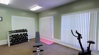 This single-story fitness space offers a functional and welcoming environment for exercise and wellness activities. The room is finished with soft sage green walls that create a calm, motivating atmosphere, complemented by neutral carpet flooring underfoot. Two generously sized windows dressed with white vertical blinds line the walls, allowing occupants to control the amount of natural light entering the space while maintaining privacy. Overhead, a fluorescent ceiling fixture provides consistent, bright artificial lighting. The room is equipped with a dumbbell rack stocked with a range of free weights, an adjustable workout bench, a stationary exercise bike, and a yoga mat, making it suitable for a variety of fitness routines including strength training, cardio, and flexibility work. White baseboards provide clean trim detail throughout, and the overall layout offers enough open floor space to move freely between equipment. The space is practical and straightforward in design, prioritizing functionality while maintaining a tidy, organized appearance ideal for daily workouts.