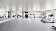 Located at 1402 Tacoma Ave S in Tacoma, Washington, this well-appointed amenity space spans a generous single floor, thoughtfully designed to serve both fitness and collaborative needs. The layout encompasses distinct zones, each with its own character and purpose.

The fitness center is the centerpiece of the space, bathed in natural light streaming through a series of large windows that line the perimeter. The open, rubber-floored exercise area houses an impressive array of equipment, including treadmills, stationary bikes, a cable weight machine, free weights with a dedicated rack, and stability balls. Floor-to-ceiling mirrors amplify the sense of space, while the clean white ceiling and bright, airy atmosphere create an energizing workout environment with climate control overhead.

Adjacent to the fitness area, a sophisticated lounge area provides a welcoming contrast, featuring exposed concrete accent walls, curated artwork, and plush rounded seating arranged on a layered geometric rug. Tall potted trees add a natural, organic element against the light-filled windows.

The conference and meeting room offers a polished, functional setting anchored by a solid wood communal table illuminated by track lighting. A dramatic full-wall photographic mural depicting a serene mountain lake scene serves as a striking focal point, complemented by a bold yellow accent rug and warm-toned furnishings. A reception or lobby area with a geometric feature wall in sage green ties the entire space together with modern architectural detail.
