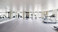 Located at 1402 Tacoma Ave S in Tacoma, Washington, this well-appointed amenity space spans a generous single floor, thoughtfully designed to serve both fitness and collaborative needs. The layout encompasses distinct zones, each with its own character and purpose.

The fitness center is the centerpiece of the space, bathed in natural light streaming through a series of large windows that line the perimeter. The open, rubber-floored exercise area houses an impressive array of equipment, including treadmills, stationary bikes, a cable weight machine, free weights with a dedicated rack, and stability balls. Floor-to-ceiling mirrors amplify the sense of space, while the clean white ceiling and bright, airy atmosphere create an energizing workout environment with climate control overhead.

Adjacent to the fitness area, a sophisticated lounge area provides a welcoming contrast, featuring exposed concrete accent walls, curated artwork, and plush rounded seating arranged on a layered geometric rug. Tall potted trees add a natural, organic element against the light-filled windows.

The conference and meeting room offers a polished, functional setting anchored by a solid wood communal table illuminated by track lighting. A dramatic full-wall photographic mural depicting a serene mountain lake scene serves as a striking focal point, complemented by a bold yellow accent rug and warm-toned furnishings. A reception or lobby area with a geometric feature wall in sage green ties the entire space together with modern architectural detail.