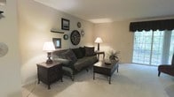 This single-floor residence offers a warm and inviting living space defined by a classic, understated aesthetic. The living room serves as the heart of the home, featuring neutral beige walls and plush, light-colored carpeting that together create a calm and cohesive atmosphere. A large window allows natural light to flow into the space, brightening the room and offering a connection to the outdoors.

The living room is thoughtfully furnished with a substantial upholstered sofa in deep, rich tones, complemented by decorative throw pillows. Dark wood accent tables flank the seating area, each topped with elegant table lamps that provide a warm, ambient glow. A sturdy dark wood coffee table anchors the center of the space, adding a sense of grounded sophistication.

The walls are tastefully decorated with an artful arrangement of wall clocks, framed artwork, and a decorative sunburst mirror, giving the room a curated, lived-in character. A small decorative shelf adds additional display space for personal accents. The overall ambiance of the space is one of timeless comfort — refined yet approachable, with carefully chosen furnishings and décor that reflect a cohesive, classic design sensibility throughout.