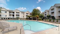 This residential community centers around a standout outdoor amenity area highlighted by a large, sparkling swimming pool with gently curved edges and a clean tiled surround. The pool deck is furnished with multiple lounge chairs and seating arrangements, providing ample space for relaxation and recreation under open skies. A vine-covered pergola structure anchors one side of the pool area, offering a shaded gathering spot equipped with additional seating and a grill, making it well-suited for outdoor entertaining. Mature trees and neatly trimmed hedgerows frame the perimeter, adding greenery and a sense of privacy. A decorative iron fence encloses the pool area, while the multi-story residential buildings surrounding the courtyard feature neutral beige exteriors, pitched rooflines, and private balconies that overlook the pool below.

The interior includes a dedicated office or utility room finished with warm hardwood flooring, crown molding, and a two-tone wall treatment featuring a deep navy lower half and taupe upper walls. A tall window dressed with horizontal blinds allows natural light into the space. The room's clean, classic details give it a polished and functional character suited for administrative use.