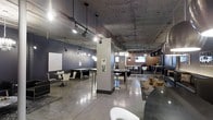 This single-floor commercial space showcases a distinctive industrial aesthetic, blending raw architectural elements with modern design sensibilities. The exposed concrete ceiling commands attention overhead, fitted with a combination of track lighting and sleek pendant fixtures that cast warm, focused light across the open floor plan. Polished concrete floors run throughout, adding to the space's urban character while remaining highly functional.

The layout is thoughtfully organized into multiple seating zones, offering a variety of configurations to accommodate different uses. High-top bar tables with chrome stools provide casual perching spots, while lower seating areas with contemporary chairs and small tables create more intimate settings. Structural columns punctuate the open floor plan, subtly defining zones without enclosing the space.

Dark, moody wall tones contrast with bright pendant globe lights and metallic accents, contributing to an atmosphere that is both energetic and refined. A display screen mounted on a warm wood-paneled accent wall adds a functional focal point on one end of the space. The overall ambiance is dynamic and versatile, making this space well-suited for a lounge, co-working environment, or collaborative gathering venue.