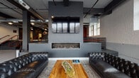 This single-floor space masterfully blends industrial heritage with contemporary design. The main entrance opens through a striking wall of multi-pane glass doors, revealing polished concrete floors and exposed structural columns that speak to the building's original character. White-painted brick walls line the interior, complemented by dark-painted ceilings through which exposed ductwork and utility pipes run overhead, reinforcing the industrial aesthetic throughout.

At the heart of the space lies a sophisticated lounge area anchored by a sleek, wall-mounted linear fireplace paired with a large flat-screen TV above. Tufted black leather Chesterfield sofas flank a live-edge wood coffee table adorned with natural succulents, creating an inviting and curated atmosphere. Tall, multi-pane arched windows allow abundant natural light to pour in, enhancing the open, airy feel of the room.

A dramatic staircase with dark treads and bold red-orange railings connects to an upper mezzanine level, serving as a striking architectural focal point. The space also features a corridor with dual stainless steel elevators, ensuring accessibility throughout. Pendant and industrial-style ceiling fixtures provide warm accent lighting, while curated artwork displayed on the walls adds a gallery-like quality. The overall ambiance is sophisticated, urban, and versatile.