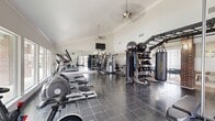 This single-level fitness space offers a thoughtfully designed environment for a full-body workout experience. The room is defined by its vaulted ceilings, which lend an open, airy quality to the interior, and are complemented by pendant-mounted ceiling fans that ensure comfortable airflow throughout. Crown molding adds a refined architectural detail along the upper walls, while large windows along the perimeter flood the space with natural light and provide views of the surrounding exterior.

The flooring consists of dark, polished slate-style tiles that extend throughout the entire area, contributing to a sleek and functional aesthetic. Full-length mirrors are positioned along one wall, visually expanding the space and serving as a practical training aid. A wall-mounted flat-screen television provides entertainment during workouts.

The equipment layout is organized and comprehensive. A dedicated cardio zone lines one side of the room, featuring multiple elliptical trainers, treadmills, and rowing machines. The strength training area includes a power rack with an adjustable bench, a multi-station cable machine, a full dumbbell rack, kettlebell sets, medicine balls, resistance bands, and a heavy punching bag. A brick accent column adds a subtle architectural contrast to the otherwise neutral white walls. A separate entry door connects to an adjoining space.