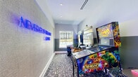Located in Huntsville, Texas, this dedicated arcade gaming room is a compact yet energetic entertainment space packed with personality. The room's two-tone walls — light gray on top and a deeper charcoal on the bottom — provide a sleek, modern backdrop that complements the vivid neon lighting and colorful arcade machines throughout. A patterned carpet in muted tones lines the floor, adding texture beneath the lively array of equipment.

The space is outfitted with an impressive collection of arcade and gaming machines, including an Avengers-themed pinball machine, a Big Buck Hunter: Reloaded shooting arcade, a Golden Tee PGA golf simulator, and a Pac-Man Battle Royale Chomp Mania tabletop unit complete with swivel stools. Bold neon signage on the wall reinforces the non-stop gaming atmosphere.

Natural light enters through a large window that frames a view of a resort-style swimming pool and surrounding residential buildings, offering a pleasant contrast to the neon-lit interior. A glass-paneled door provides access to an adjacent space, keeping the room open and connected. Recessed ceiling lighting ensures the space is well-lit, while the glowing blue LED accents on several machines add to the immersive, arcade-hall ambiance. This room delivers a concentrated, high-energy gaming experience.