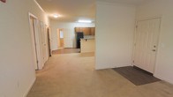 This single-story space presents an open-concept layout that seamlessly connects the main living area to the kitchen. The living space is finished with wall-to-wall beige carpeting, contributing to a warm and comfortable atmosphere, while an adjacent area features dark hardwood-style flooring, providing a subtle contrast in material and texture throughout the unit.

The kitchen is positioned toward the rear of the open space and is equipped with medium-toned wood cabinetry, a black refrigerator, and a peninsula-style countertop that serves as a natural divider between the kitchen and the living area. This layout supports both functionality and an open flow between spaces.

Crown molding accents the ceiling, adding a refined architectural detail to the otherwise understated interior. Flush-mounted ceiling lighting provides consistent illumination throughout. A hallway leads to additional rooms, including what appears to be a bedroom with a window allowing natural light. Multiple white panel doors are visible, indicating access to several separate rooms. The overall palette of neutral whites and beiges creates a clean, move-in ready environment.