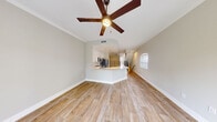 This two-story home presents a clean, modern aesthetic anchored by warm wood-look flooring that runs throughout the main level. The open-concept living area is bright and airy, enhanced by crown molding, neutral walls, and a ceiling fan overhead. A central kitchen sits at the heart of the main floor, elevated slightly and open to the living space below. It features white shaker-style cabinetry, a white subway tile backsplash, stainless steel appliances including a refrigerator, range, and microwave, as well as a generous quartz-topped peninsula ideal for dining or food preparation. Recessed lighting illuminates the kitchen, complementing the natural light that filters in through shuttered windows.

A sleek staircase with wood-look treads, white risers, and a modern black metal railing connects the two floors, adding a contemporary architectural detail. The upper level transitions to plush carpeted bedrooms finished in soft neutral tones, each equipped with ceiling fans. The bedrooms are well-proportioned and bright, with access to a hallway bathroom featuring tiled walls and quartz countertops. Throughout, the design maintains a consistent, move-in-ready aesthetic with clean finishes and a cohesive neutral palette.