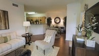 This single-floor residence is designed around an open-concept floor plan that integrates the living, dining, and kitchen areas into one cohesive, flowing space. Warm hardwood flooring extends throughout, lending a sense of continuity and elegance to the interior.

The living area serves as the central gathering space, highlighted by a tufted accent chair and a patterned area rug that anchors the room. A round side table with a classic lamp adds a refined decorative touch. The space transitions naturally into the dining area, which features a round dining table surrounded by upholstered chairs, accented by a decorative mirror and flanking potted trees that introduce a natural, organic element.

Adjacent to the dining area, the kitchen is partially open to the main living space via a raised counter pass-through, providing both functional utility and visual connection. The kitchen features warm wood cabinetry and overhead lighting. Crown molding details along the upper walls contribute a polished architectural finish. The overall ambiance is warm, inviting, and well-appointed, reflecting a thoughtfully designed single-level layout.