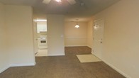 This single-floor space is designed with an open, interconnected layout that promotes ease of movement between its main areas. The living area features wall-to-wall carpet in a neutral brown tone, providing a comfortable and cohesive foundation underfoot. A ceiling fan is mounted overhead, offering both lighting and air circulation to the space.

Adjacent to the living area is a defined dining nook, distinguished by a raised platform and illuminated by a hanging pendant light that serves as a modest focal point. The subtle separation created by the step-up platform gives the dining area its own identity while maintaining visual continuity with the rest of the space.

The kitchen is positioned off to the side, finished with light-toned cabinetry and white appliances, including a freestanding electric range. The kitchen benefits from its own overhead fluorescent lighting, keeping the workspace bright and functional. White trim and baseboards run consistently throughout, complementing the beige walls and contributing to a clean, uniform aesthetic. The front entry door is visible from the main living area, reinforcing the compact and efficiently organized nature of this single-level space.