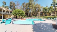 Located at 444-445 N Amelia Ave in San Dimas, California, this single-level outdoor amenity space offers a well-appointed community environment designed for relaxation and recreation. At the heart of the space is a generously sized resort-style swimming pool with a connected spa, surrounded by a spacious concrete deck furnished with colorful lounge chairs and seating areas. A standout natural feature is a dramatic rock waterfall installation, where cascading water flows over stacked boulders into a shallow grotto pool, framed by lush tropical foliage including banana plants, palms, and dense greenery that creates a secluded, garden-like atmosphere. Adjacent to the pool area, a shaded cabana pavilion provides an elegant outdoor lounge space outfitted with wicker sofas and accent chairs accented in navy and orange, draped with navy curtains for privacy and shade. Curved pathways wind through manicured lawns, colorful flower beds, and mature trees, connecting the various outdoor areas and lending a park-like quality to the grounds. The overall ambiance is bright, tropical, and resort-inspired, with the surrounding two-story residential buildings visible in the backdrop, well-integrated into the verdant landscape under the sunny Southern California sky.