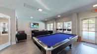 This single-floor community clubhouse is thoughtfully designed to serve as a social and recreational hub. The main gathering area is anchored by a striking blue-felt pool table at its center, complemented by sleek hardwood-style flooring, recessed and flush-mount lighting, and a wall-mounted flat-screen television. Comfortable black leather seating — including a sofa and accent chairs — creates an inviting lounge environment, while large windows along the perimeter flood the space with natural light and offer views of the surrounding outdoor pool area and landscaping.

A dedicated lounge nook provides a more intimate setting with additional seating, wall art, and a cozy atmosphere for relaxed gatherings. Adjacent to the main area, a kitchenette features gray cabinetry, light stone-look countertops, a double basin sink, and ample storage — ideal for hosting events and communal use.

A double glass-door entry leads into the fitness center, which is visible through the panes and equipped with cardio machines set on durable flooring. The entryway features a rich plum-toned accent wall and elegant white decorative vases, setting a polished tone upon arrival. Throughout, the neutral color palette, wood-look flooring, and generous natural light create a bright, modern, and welcoming atmosphere suited for both active recreation and casual socializing.