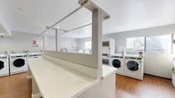 Located at 150 W Hill Ave in Fullerton, California, this shared laundry facility offers a clean, functional, and efficiently organized space designed to serve the needs of its users. The room features warm hardwood-style flooring that contrasts neatly with the light gray and white walls, giving the space a tidy and welcoming feel. Recessed ceiling lighting provides consistent, even illumination throughout the room.

The facility is well-stocked with a substantial number of front-loading washing machines and stacked dryer units, all finished in white and arranged along the walls to maximize usable floor space. A long, white folding counter runs along the center of the room, supported by a structural divider, offering ample workspace for sorting and folding laundry. Additional top-loading washers are also available along one wall.

A snack and beverage vending machine is conveniently situated near the entrance, adding an extra layer of convenience for users during longer laundry sessions. Informational and instructional signage is posted on the walls, helping users navigate available services and payment options. Natural light filters in through windows on multiple sides of the room, complementing the recessed lighting for a bright overall atmosphere. The space is thoughtfully laid out to accommodate multiple users simultaneously with ease.
