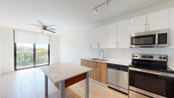 This single-floor unit features a contemporary, open-concept design with light wood-look tile flooring throughout, creating a cohesive and airy feel across all spaces. The kitchen serves as a functional centerpiece, showcasing a two-tone cabinetry scheme that pairs high-gloss white upper cabinets with warm wood-grain lower cabinets. A textured tile backsplash adds visual interest, while a full suite of stainless steel appliances — including a refrigerator with ice maker, electric range, over-the-range microwave, and dishwasher — provides modern convenience. A freestanding island with a white quartz-style top and wood shelf below offers additional workspace and casual dining. Track lighting and recessed fixtures illuminate the kitchen effectively. The living area flows naturally from the kitchen, featuring a ceiling fan and access to a window with outdoor views. The bedroom is a bright, serene space with its own ceiling fan, clean white walls, and a window that invites natural light and treetop views. The bathroom is finished with large-format wood-look wall tiles in the tub/shower enclosure, a soaking tub, a rain-style showerhead, and matching floor tiles, creating a spa-like atmosphere. Throughout, the unit maintains a clean, minimalist aesthetic.