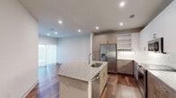 This contemporary single-floor residence showcases a thoughtfully designed open-concept layout that seamlessly connects the kitchen and living areas. The kitchen is a standout centerpiece, featuring a large island with granite countertops and an undermount sink, complemented by two-tone cabinetry in warm taupe and crisp white. Stainless steel appliances—including a French door refrigerator with ice maker, a slide-in range, a built-in microwave, and a dishwasher—provide a sleek, modern finish. Rich, dark hardwood flooring runs throughout the main living areas, adding warmth and sophistication, while recessed lighting illuminates the space from above. Open shelving and ample cabinetry offer generous storage throughout the kitchen. The bedroom is a comfortable retreat, carpeted for softness and equipped with a ceiling fan and a window that brings in natural light. The bathroom is appointed with large-format white tile flooring, a dual-sink vanity topped with granite, warm wood cabinetry, and a broad frameless mirror. A walk-in closet accessible directly from the bathroom provides excellent storage. Overall, the residence presents a clean, modern aesthetic with quality finishes and a practical, open flow throughout.