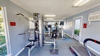 Located at 6184 Cottle Road in San Jose, California, this fitness center offers a compact yet well-equipped exercise space designed to support a well-rounded workout routine. The room is finished with soft blue-gray walls, a textured ceiling, and a patterned low-pile carpet that adds visual interest underfoot while providing a practical workout surface.

The space is outfitted with a variety of cardio and strength training equipment, including a treadmill, elliptical machine, stationary bike, rowing machine, and a multi-station cable weight machine with an adjustable bench. Full-length mirrors span one wall, creating a sense of added depth and allowing users to monitor their form during exercise.

Natural light flows in through large windows and a glass-paneled entry door, offering pleasant views of the surrounding landscaped outdoor area with mature flowering trees. Fluorescent overhead lighting ensures the space remains bright throughout the day. A wall-mounted air conditioning unit provides climate control for year-round comfort. A wall-mounted defibrillator and safety signage reflect attention to occupant safety. The fitness center is clearly identified by signage on the entry door and is designed to serve as a convenient, functional amenity for the surrounding community.