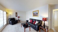 This single-floor residence in Williamsburg offers a well-appointed and comfortable living environment designed for everyday ease. The living room serves as the heart of the home, showcasing plush wall-to-wall carpeting that adds warmth underfoot and a soft, neutral color palette throughout. The textured ceiling and clean white walls provide a classic backdrop, allowing furnishings and décor to stand out effortlessly. The space is generously proportioned, accommodating a full seating arrangement with ample room to move freely. A centrally placed sofa with accent pillows anchors the room, complemented by coordinating seating and wooden end tables that add a touch of traditional charm. Decorative lamps cast a warm, inviting glow, while framed wall art lends a personalized character to the space. A closet door visible along one wall suggests convenient built-in storage. The overall ambiance of this residence is cozy and livable, with a straightforward, functional layout that makes efficient use of every area. The neutral tones and classic finishes throughout create a versatile canvas well-suited to a variety of interior styles and personal tastes.