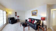 This single-floor residence in Williamsburg offers a well-appointed and comfortable living environment designed for everyday ease. The living room serves as the heart of the home, showcasing plush wall-to-wall carpeting that adds warmth underfoot and a soft, neutral color palette throughout. The textured ceiling and clean white walls provide a classic backdrop, allowing furnishings and décor to stand out effortlessly. The space is generously proportioned, accommodating a full seating arrangement with ample room to move freely. A centrally placed sofa with accent pillows anchors the room, complemented by coordinating seating and wooden end tables that add a touch of traditional charm. Decorative lamps cast a warm, inviting glow, while framed wall art lends a personalized character to the space. A closet door visible along one wall suggests convenient built-in storage. The overall ambiance of this residence is cozy and livable, with a straightforward, functional layout that makes efficient use of every area. The neutral tones and classic finishes throughout create a versatile canvas well-suited to a variety of interior styles and personal tastes.