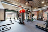 Located in Charlotte, NC, this fitness center combines industrial design with a comprehensive workout environment. The space is defined by its striking reclaimed wood plank ceiling, exposed silver ductwork, and large ceiling fans that contribute to an open, loft-like atmosphere. Structural steel columns punctuate the floor plan, reinforcing the industrial aesthetic throughout.

The exercise room is outfitted with a diverse range of professional-grade equipment to support varied training styles. Cardio offerings include AirRaid curved treadmills and air resistance bikes, while a central multi-functional rig accommodates TRX suspension training, resistance bands, and a punching bag. Rowing machines are lined along one side of the space, and a dedicated strength area features a squat rack with an extensive collection of color-coded bumper plates, kettlebells, medicine balls, and stability balls organized on storage racks.

Large-format steel-framed glass partitions run along one wall, flooding the interior with natural light and providing views into adjacent areas. A full-length mirror expands the visual depth of the room. On one end, a roll-up garage-style door opens directly to an outdoor patio, offering fresh air and seamless indoor-outdoor connectivity. An artistic, colorful accent wall adds a bold visual focal point, complemented by exposed brick detailing. Durable, low-pile carpet tiles cover the floor throughout, providing a practical surface suited to high-intensity workouts.