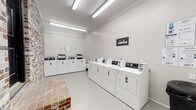 Located at 4827 Jersey Pike in Chattanooga, Tennessee, this single-floor laundry facility is designed for efficient, high-capacity use. The room is outfitted with a full lineup of commercial-grade top-load washing machines and front-load dryers arranged along the walls, providing ample capacity for multiple users simultaneously. The space is finished with neutral gray walls and a speckled hard-surface floor that is easy to maintain and keep clean. Overhead fluorescent lighting runs the length of the ceiling, ensuring the room is well-lit throughout. A notable design feature is the exposed brick wall along one side of the room, adding a touch of character and texture that contrasts with the otherwise clean, modern aesthetic. A brick seating or folding surface is also present near the entrance, offering a practical spot for users. Branded signage for "Laundry Again" is displayed on the wall, indicating the facility's service affiliation. The overall layout is open and organized, maximizing accessibility and ease of movement around the equipment. This facility provides a well-maintained, functional environment suited to serve the laundry needs of a residential community or similar setting.
