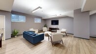 Located at 208 Woolwich St S, this thoughtfully designed amenity space offers a versatile, single-level layout that seamlessly blends social, recreational, and functional areas. The main lounge area is anchored by a bold blue sofa and a collection of mid-century modern armchairs arranged around a round coffee table, all set atop light hardwood-style flooring. A wall-mounted flat-screen TV above an electric fireplace serves as the focal point of this relaxing gathering space, while linear pendant lighting illuminates the room from above. Natural light filters in through high horizontal windows, lending an airy feel to the interior.

The meeting and activity area features multiple dark wood tables paired with grey upholstered chairs, glass whiteboard panels mounted on the walls, and framed notices — making it well-suited for both collaborative work and casual games, as evidenced by the chess set on one of the tables.

A compact kitchenette rounds out the space, finished with crisp white shaker-style cabinetry, a stainless steel sink, dishwasher, full-size refrigerator, microwave, toaster oven, and a coffee station. The clean, neutral grey tones throughout create a cohesive and modern aesthetic, making this space functional and welcoming for a variety of uses.