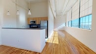 Unit 603 is a one-bedroom residence with a distinctive loft-inspired design that blends industrial character with modern finishes. The open-concept layout is anchored by soaring ceilings with exposed ductwork painted white, giving the space an airy, contemporary feel. Large, grid-paned industrial windows line the main living area, flooding the hardwood floors with abundant natural light and offering views of the surrounding cityscape.

The kitchen is integrated seamlessly into the living space via a peninsula with a dark countertop, providing both a functional workspace and a natural room divider. Light maple cabinetry pairs with stainless steel appliances—including a refrigerator, electric range, microwave, and dishwasher—while a checkered tile floor adds a classic accent underfoot. Pendant lighting above the peninsula adds a focused, stylish touch.

The bedroom is generously proportioned, benefiting from the same large windows and hardwood flooring found throughout. Natural wood doors add warmth to the otherwise crisp, neutral palette. The bathroom features classic black-and-white mosaic tile flooring, a full bathtub with shower, and maple vanity cabinetry, completing the unit's cohesive mix of industrial and refined residential design.