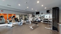 This fitness center is designed to support a full range of workout routines within a spacious, single-floor layout. The space is divided into distinct exercise zones, each thoughtfully arranged to maximize functionality. The cardio area features multiple treadmills and elliptical trainers positioned near large windows that allow natural light to fill the room, creating an energizing atmosphere for exercise. A dedicated strength training section includes a cable machine and an adjustable weight bench, offering versatility for resistance-based workouts. A well-stocked dumbbell rack lines one wall, providing an organized free weights area ideal for targeted exercises. Large wall-to-wall mirrors run along multiple walls, visually expanding the space while allowing users to monitor their form. The flooring throughout consists of durable rubber gym matting, providing both safety and comfort underfoot. Recessed lighting ensures consistent, bright illumination across the entire floor. Wall-mounted flat-screen televisions are positioned for easy viewing from various workout stations. Vibrant orange accent walls in the connecting area add a bold, motivating pop of color, complementing the cool gray tones that dominate the space. The overall ambiance is clean, modern, and purpose-built for an effective fitness experience.