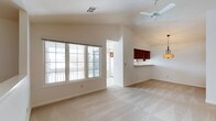 This single-floor residence offers an open and airy layout designed for comfortable everyday living. The main living area immediately draws the eye upward with its vaulted ceiling, which adds a sense of generous vertical space and enhances the room's overall brightness. A ceiling fan is centrally mounted, providing both practical airflow and a clean, understated focal point overhead. Soft, neutral-toned carpeting covers the living area floor, lending warmth and a welcoming feel underfoot. Natural light pours in through a large multi-pane window that frames views of the surrounding neighborhood, creating a bright and inviting atmosphere throughout the day. The open floor plan flows naturally into the kitchen area, which features rich, dark-toned cabinetry above a peninsula-style counter — ideal for casual dining or meal prep. A pendant light fixture adds a touch of character to this transitional space. The crisp white walls throughout maintain a clean, neutral palette that complements the warm wood tones of the cabinetry. A hallway visible from the main living space suggests additional rooms beyond, rounding out this well-connected and functional single-level home.