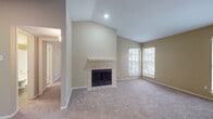 This single-floor residence is defined by its vaulted ceilings in the living area, which create a dramatic sense of height and openness. The living room serves as the centerpiece of the home, anchored by a tile-surround fireplace with a clean white mantle and finished with plush carpeting underfoot. Multiple windows along the living room walls flood the space with natural light and offer views of surrounding greenery, enhancing the bright, welcoming atmosphere.

The layout transitions seamlessly into an open kitchen, visible through a pass-through opening from the living area. The kitchen is well-appointed with dark espresso cabinetry reaching toward the ceiling, light granite-style countertops, wood-look plank flooring, and a full suite of black appliances including a range, microwave, refrigerator, and dishwasher. A peninsula provides additional prep and serving space.

The bedrooms are carpeted and finished in warm neutral tones, each featuring a chair-rail detail and access to closets with built-in shelving systems for organized storage. One bedroom connects directly to a bathroom featuring dark vanity cabinetry and light countertops. Throughout the home, warm beige walls and consistent finishes create a cohesive, functional living environment.