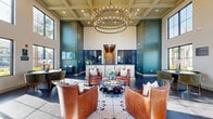 Located at 4820 SW 48th Ave in Ocala, Florida, this elegantly appointed single-floor space serves as a refined lobby and lounge area. The defining architectural feature is its dramatic double-height ceiling, which creates an expansive, airy atmosphere and is anchored by a striking two-tiered circular chandelier adorned with candelabra-style bulbs. A coffered ceiling detail above the central seating area adds a layer of architectural sophistication.

The flooring transitions between large-format dark marble-effect tiles and a decorative mosaic tile inlay beneath the central seating arrangement, contributing both visual interest and definition to the space. Floor-to-ceiling windows along multiple walls flood the interior with abundant natural light and provide open views of the surrounding landscaping and outdoor areas.

The main lounge area is furnished with rich cognac leather swivel chairs arranged around a sleek round marble-top coffee table, creating an inviting conversational setting. A separate dining nook features a round wood table paired with tufted green velvet chairs. Deep teal accent walls frame a reception counter area, adding bold color contrast to the neutral beige walls. Open metal-and-wood shelving units serve as stylish display and storage elements. The overall ambiance is polished, warm, and professionally designed — ideal for welcoming guests.