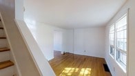This multi-level property spans three floors, offering a well-structured layout suited for a variety of living arrangements. The interior is defined by warm hardwood flooring that runs throughout the main living areas, lending a timeless and classic character to the space. Walls are finished in crisp, clean white throughout, creating a bright and neutral backdrop that allows for flexible interior styling and décor.

The main living area is open and airy, bathed in natural light that filters in and casts warm patches across the hardwood floors. The room flows naturally into an adjacent space through a wide doorway, suggesting a connected and functional floor plan. Ceiling heights are standard, contributing to a comfortable and approachable atmosphere. Baseboard details and floor vents are visible, indicating a well-maintained infrastructure throughout.

The staircase, visible from the main level, provides access between the three floors, each of which likely offers additional living or utility space. The overall aesthetic is clean and understated, with a focus on practical design and durable materials. The white walls and natural wood floors combine to create a cohesive, inviting environment that serves as an excellent canvas for personalization.