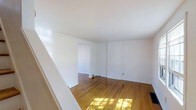 This multi-level property spans three floors, offering a well-structured layout suited for a variety of living arrangements. The interior is defined by warm hardwood flooring that runs throughout the main living areas, lending a timeless and classic character to the space. Walls are finished in crisp, clean white throughout, creating a bright and neutral backdrop that allows for flexible interior styling and décor.

The main living area is open and airy, bathed in natural light that filters in and casts warm patches across the hardwood floors. The room flows naturally into an adjacent space through a wide doorway, suggesting a connected and functional floor plan. Ceiling heights are standard, contributing to a comfortable and approachable atmosphere. Baseboard details and floor vents are visible, indicating a well-maintained infrastructure throughout.

The staircase, visible from the main level, provides access between the three floors, each of which likely offers additional living or utility space. The overall aesthetic is clean and understated, with a focus on practical design and durable materials. The white walls and natural wood floors combine to create a cohesive, inviting environment that serves as an excellent canvas for personalization.