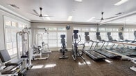 This fitness center offers a comprehensive and well-organized workout environment designed to support a wide range of exercise routines. The space is filled with an impressive array of cardio equipment, including multiple treadmills and elliptical trainers, as well as stationary bikes positioned throughout the room. Strength training is well represented with cable machines, weight benches, medicine ball racks, and resistance equipment available for full-body workouts.

Architecturally, the facility features crown molding, decorative columns, and a tray ceiling with recessed skylight panels that provide bright, even overhead lighting. Multiple ceiling fans enhance air circulation throughout the space. A wall of floor-to-ceiling mirrors runs along one side of the room, visually expanding the area and including a classic ballet barre — a thoughtful touch for stretching or dance-based exercise.

Large, multi-pane windows fitted with white plantation shutters line the exterior walls, drawing in generous natural light and offering views of the surrounding greenery. The textured, patterned carpet flooring adds a refined finish underfoot. A glazed entry door with sidelights provides an elegant point of entry. The overall ambiance is clean, bright, and inviting — a polished fitness space suited for a variety of training styles.
