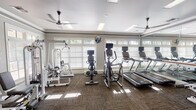 This fitness center offers a comprehensive and well-organized workout environment designed to support a wide range of exercise routines. The space is filled with an impressive array of cardio equipment, including multiple treadmills and elliptical trainers, as well as stationary bikes positioned throughout the room. Strength training is well represented with cable machines, weight benches, medicine ball racks, and resistance equipment available for full-body workouts.

Architecturally, the facility features crown molding, decorative columns, and a tray ceiling with recessed skylight panels that provide bright, even overhead lighting. Multiple ceiling fans enhance air circulation throughout the space. A wall of floor-to-ceiling mirrors runs along one side of the room, visually expanding the area and including a classic ballet barre — a thoughtful touch for stretching or dance-based exercise.

Large, multi-pane windows fitted with white plantation shutters line the exterior walls, drawing in generous natural light and offering views of the surrounding greenery. The textured, patterned carpet flooring adds a refined finish underfoot. A glazed entry door with sidelights provides an elegant point of entry. The overall ambiance is clean, bright, and inviting — a polished fitness space suited for a variety of training styles.