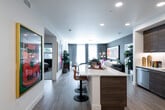This thoughtfully designed single-floor residence combines contemporary style with bold, expressive interiors. The open-concept living and kitchen area serves as the heart of the home, anchored by a sleek island with a white quartz countertop and warm wood-toned base, complemented by adjustable bar seating. The kitchen is outfitted with full-height dark wood cabinetry, stainless steel appliances including a gas range and dishwasher, and a chevron-patterned tile backsplash. A teal accent wall adds a striking visual focal point to the open living space, which flows seamlessly into a bright, windowed sitting area adorned with curated artwork and indoor greenery. The living room features a mid-century modern media console, large-scale wall art, and direct access to an outdoor balcony, allowing natural light to fill the space. The bedroom is a serene retreat with generous window coverage, a bold upholstered headboard, and warm wood furnishings. Two full bathrooms each make a design statement — one with deep navy walls, a backlit vanity mirror, walk-in shower, and access to a walk-in closet, and the other with vivid magenta walls, tiled tub surround, and a floating wood vanity. Light wood-tone flooring runs throughout, unifying the space with a clean, modern aesthetic.