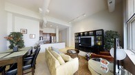 This thoughtfully designed single-floor residence offers a seamless open-concept layout that connects the living, dining, and kitchen areas into one cohesive and inviting space. The living room is anchored by a plush, cream-toned sectional sofa paired with a round coffee table and a large area rug, creating a warm and comfortable gathering area. A substantial dark-finished entertainment unit with display shelving lines one wall, providing both storage and a striking focal point. Adjacent to the living space, a dining nook with sleek dark chairs offers an intimate setting for meals. The kitchen features rich dark wood cabinetry, stainless steel appliances including a refrigerator, and a breakfast bar peninsula that opens directly to the living area, enhancing the sense of flow and connectivity. Soaring ceilings fitted with track lighting illuminate the entire space efficiently, while recessed lighting adds warmth. The neutral color palette of whites, creams, and warm tones throughout gives the residence a clean, modern aesthetic. Soft carpeting underfoot adds to the overall comfort of the living areas, making this a polished and functional space well-suited for both relaxed everyday living and entertaining.