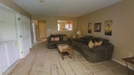 This single-story space centers around a spacious living room designed for comfort and functionality. The room is finished in warm, neutral beige tones on the walls and features wall-to-wall carpeting complemented by a large patterned area rug, adding texture and visual interest underfoot. Two substantial dark brown upholstered sofas anchor the seating area, arranged around a wooden coffee table with a glass top surface. Decorative pillows in earth tones and accent artwork on the walls contribute to the cohesive, understated aesthetic.

A notable architectural feature is the pass-through window that opens directly into the kitchen, allowing natural sightlines between the two spaces and supporting an open, connected feel. The kitchen, visible through the opening, features warm wood cabinetry. A hallway extending from the living area leads toward additional rooms, with a door at the far end suggesting direct exterior access. The space receives natural light through a window along the hallway wall. A table lamp provides warm ambient lighting within the living area, enhancing the inviting atmosphere throughout.