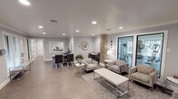 Located at 4850 River Ranch Blvd in Fort Worth, Texas, this single-story community clubhouse and leasing office delivers a polished, contemporary environment designed to welcome and serve residents. The main lounge area is an open, generously proportioned space finished with wood-look tile flooring, neutral gray walls, and recessed lighting that creates a bright and inviting atmosphere. Comfortable patterned accent chairs and sofas are arranged around a central coffee table, anchored by a decorative area rug, while a distinctive exposed brick column adds a touch of architectural character and warmth. An adjacent fitness center is visible through large glass-paned sliding doors, equipped with cardio machines and offering natural light from surrounding windows. The leasing office area features a dedicated workstation with dual monitors, a service counter pass-through window, and additional seating along windowed walls that fill the space with natural light. A separate workspace zone includes a sleek round dining-style table with white chairs set beneath vaulted ceilings, alongside a computer desk, making it ideal for resident use or private meetings. Throughout, the design maintains a cohesive modern aesthetic with chrome accents, decorative mirrors, and clean architectural lines that together create a professional yet comfortable community hub.