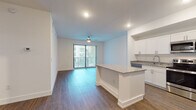 This well-appointed single-floor residence showcases a clean, contemporary design throughout. The open-concept layout seamlessly connects the kitchen and living area, creating a bright and functional shared space. The kitchen is a standout feature, equipped with stainless steel appliances including a refrigerator with ice maker, range, microwave, and dishwasher. White shaker-style cabinetry pairs elegantly with granite countertops, while a center island provides additional prep and casual dining space. Recessed lighting illuminates the area, complementing the warm hardwood-style flooring that extends throughout the unit. The living area is enhanced by a ceiling fan and offers access to a private balcony through sliding glass doors dressed with vertical blinds, inviting natural light and outdoor views. The bedrooms feature tray ceilings, adding an architectural touch, and are finished with the same rich flooring. The bathroom is outfitted with a tiled walk-in shower with glass sliding doors, a granite vanity, and modern fixtures. A dedicated laundry area houses a full-size stacked washer and dryer alongside wire shelving for added storage. Clean white walls and consistent finishes throughout lend the space a cohesive, move-in ready appeal.