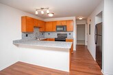 This single-floor unit at 1900 River Rd in Saint Clair, Michigan offers a well-designed, open layout with a cohesive mix of finishes throughout. The kitchen is a functional centerpiece, featuring warm honey-toned cabinetry, granite countertops, a speckled tile backsplash, and a full suite of black appliances including a range, microwave, dishwasher, and refrigerator. A breakfast bar peninsula extends into the adjoining dining or living area, providing additional counter space and a natural transition between rooms. The main living area is finished with rich hardwood-style flooring and benefits from abundant natural light through large sliding glass doors fitted with vertical blinds, creating a bright, open atmosphere. The bedrooms are carpeted and neutrally painted, offering a comfortable, quiet feel. One bedroom features a spacious walk-in closet with built-in rod storage, while another includes a standard reach-in closet. Windows in each bedroom allow for natural light. The bathroom is finished with large-format tile flooring, warm wood vanity cabinetry, granite countertops, and a soaking tub with a shower. The overall design is clean and consistent, with neutral tones throughout.