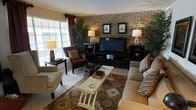 This single-floor space exudes a warm and inviting character, anchored by a striking exposed brick accent wall that serves as the centerpiece of the living area. The textured brickwork adds rustic charm and depth, providing a rich backdrop for the entertainment setup, complete with a large flat-screen TV flanked by dark wood media consoles adorned with table lamps that cast a soft, ambient glow.

The living area is thoughtfully arranged with a generous sofa dressed in striped accent pillows, a classic leather armchair, and a nailhead-trimmed side chair, all oriented around a distressed white coffee table that adds a touch of vintage elegance. A patterned area rug anchors the seating arrangement, enhancing the layered, curated aesthetic.

Natural light filters through horizontal blinds on the windows, complemented by rich burgundy draping that adds a bold, dramatic accent to the room. Lush tropical houseplants are strategically placed to introduce a fresh, organic element throughout the space.

The overall ambiance is one of relaxed sophistication — a blend of rustic and traditional design elements that come together to create a space that feels both comfortable and stylishly appointed.