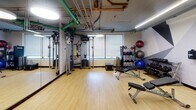 Located at 5115 N Sheridan Road in Chicago's Edgewater neighborhood, this fitness center offers a well-equipped, single-floor facility designed to support a full range of workout routines. The space is divided into distinct functional zones, each tailored to a specific type of exercise.

The strength and free weight area features light hardwood-style flooring, large wall mirrors, a Life Fitness cable machine with TRX suspension straps, adjustable weight benches, a full dumbbell rack, and an assortment of medicine and stability balls. Exposed piping overhead gives this section an industrial character.

The cardio zone transitions to dark rubber flooring and showcases a striking geometric mural in shades of grey, white, and charcoal that energizes the space. This area is outfitted with multiple Life Fitness treadmills, elliptical trainers, and upright cycling bikes, all illuminated by sleek linear LED lighting overhead.

Glass-panel partitions with black steel framing define the boundaries between zones, providing visual openness while maintaining a sense of structure. A separate enclosed area, accessible through a glass door, offers additional space for isolated strength machines or stretching.

The overall design blends industrial and modern aesthetics, with thoughtful zoning, ample equipment variety, and a functional layout that accommodates both cardio and strength training needs.