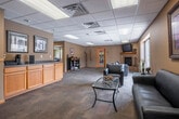 The Park Village Apartments clubhouse and leasing office is a thoughtfully designed single-floor community space offering a comfortable and functional environment for residents and visitors alike. The space is organized across two primary areas, each serving a distinct purpose.

Upon entry, a front reception desk with warm honey-toned wood cabinetry greets visitors, providing a professional and welcoming point of contact. The main lounge area extends generously from the reception, featuring plush seating arrangements, a tile-surround fireplace with a mounted flat-screen television, and decorative accents including framed artwork and ornamental plants. A dedicated dining nook with bistro-style table and chairs offers an additional casual gathering spot. Large floor-to-ceiling windows along one wall flood the space with natural light and offer views of the outdoor patio and pool area beyond.

The interior palette of warm tan and gold tones, accented by natural wood trim, creates an inviting and cohesive ambiance throughout. A suspended drop tile ceiling with integrated recessed lighting ensures consistent, well-distributed illumination across the space.

A separate business center room, branded prominently with the Park Village Apartments logo, provides residents with computer workstations, a printer, and ample desk space—ideal for professional tasks or everyday needs. Carpeted flooring throughout contributes to a quiet and comfortable atmosphere.