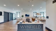 The Wynfield clubhouse at 2549 S George St in York, PA is a thoughtfully designed single-floor community amenity space that blends modern farmhouse aesthetics with functional gathering areas. Upon entry, visitors are greeted by a prominent reception desk bearing the Wynfield name, finished in a slate-blue tone that anchors the open-concept layout.

The main lounge area features warm hardwood-style flooring, crisp white walls accented by bold dark trim, and a striking tray ceiling clad in natural wood planking. A gold sputnik chandelier, wall-mounted television, and an array of comfortable seating — including tufted sofas and leather accent chairs — create an inviting atmosphere bathed in natural light from large, grid-paned windows overlooking the surrounding greenery.

Adjacent to the lounge, a well-appointed kitchen and bar area showcases dark countertops, white shaker cabinetry, a subway tile backsplash, open wood shelving, and pendant lighting above a spacious island with counter-height seating. A separate dining nook with a solid wood table, black Windsor chairs, and oversized pendant lights provides a more intimate gathering spot.

The space also includes a corridor with private rooms accessible through dark panel doors, a secondary seating lounge, and a wall-mounted display screen — offering versatile options for both social and private use throughout the community.