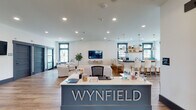 The Wynfield clubhouse at 2549 S George St in York, PA is a thoughtfully designed single-floor community amenity space that blends modern farmhouse aesthetics with functional gathering areas. Upon entry, visitors are greeted by a prominent reception desk bearing the Wynfield name, finished in a slate-blue tone that anchors the open-concept layout.

The main lounge area features warm hardwood-style flooring, crisp white walls accented by bold dark trim, and a striking tray ceiling clad in natural wood planking. A gold sputnik chandelier, wall-mounted television, and an array of comfortable seating — including tufted sofas and leather accent chairs — create an inviting atmosphere bathed in natural light from large, grid-paned windows overlooking the surrounding greenery.

Adjacent to the lounge, a well-appointed kitchen and bar area showcases dark countertops, white shaker cabinetry, a subway tile backsplash, open wood shelving, and pendant lighting above a spacious island with counter-height seating. A separate dining nook with a solid wood table, black Windsor chairs, and oversized pendant lights provides a more intimate gathering spot.

The space also includes a corridor with private rooms accessible through dark panel doors, a secondary seating lounge, and a wall-mounted display screen — offering versatile options for both social and private use throughout the community.