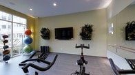 This fitness room offers a practical and inviting space for exercise, thoughtfully designed with both functionality and aesthetics in mind. The room features light-toned walls accented with mounted living wall planters filled with lush greenery, adding a natural, refreshing element to the workout environment. A large flat-screen television is mounted centrally on the main wall, accompanied by a soundbar, providing entertainment during workouts. The flooring consists of smooth, wood-style planks that are easy to maintain and contribute to a clean, modern look. Equipment visible in the space includes a stationary spin bike and a recumbent-style cardio machine, catering to a variety of cardiovascular workout preferences. Colorful exercise balls in multiple sizes are stored along the far wall, alongside rolled fitness mats, offering options for stretching and strength training. Recessed ceiling lighting provides consistent, even illumination throughout the room. The overall ambiance is energizing yet approachable, combining greenery, vibrant color accents, and well-organized equipment to create a welcoming space suited for daily fitness routines.