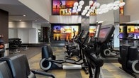 This upscale fitness center is designed to deliver a premium workout experience through thoughtful architecture and a comprehensive selection of equipment. The main cardio zone is an expansive, open area defined by soaring ceilings adorned with a dramatic cluster of spherical globe pendant lights that create a bright, energizing atmosphere. Floor-to-ceiling mirrors line the walls, visually doubling the sense of space while allowing users to monitor their form. A patterned carpet in gold and grey tones adds a sophisticated, contemporary aesthetic underfoot. The cardio floor is outfitted with a full row of Life Fitness treadmills, elliptical trainers, recumbent bikes, and upright cycles, each equipped with individual screens. A striking feature wall displays large, vibrant backlit panels depicting athletes in motion against vivid sunset backdrops, providing constant visual motivation. Transitioning into the strength training area, the flooring shifts to a durable hardwood-style surface, and the space is equipped with cable machines, Smith machines, weight benches, and a medicine ball rack. Mirrored walls continue throughout, enhancing the sense of depth. A dedicated storage and amenity station near the entrance features built-in dark cabinetry with cubbies and a granite-style countertop, offering convenient storage for personal items.