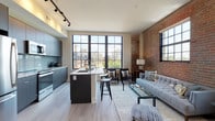 This single-floor unit showcases a compelling blend of industrial heritage and modern design. The open-concept living and kitchen area serves as the heart of the space, anchored by a dramatic exposed brick accent wall and floor-to-ceiling black-framed windows that flood the interior with natural light and offer urban views of the surrounding cityscape. Light hardwood flooring runs throughout, complementing the clean, contemporary aesthetic.

The kitchen is thoughtfully designed with sleek grey cabinetry extending to the ceiling, a tiled backsplash, stainless steel appliances including a full-size refrigerator and microwave, and a central island with a white quartz countertop and bar seating for casual dining. Track lighting overhead ensures ample illumination. The living area, bordered by the iconic brick wall, provides a relaxed yet stylish setting with generous natural light from multiple large windows.

Two bedrooms offer comfortable retreats, each featuring light hardwood floors, black-framed windows with urban views, and ample wall space. Two full bathrooms serve the unit, each equipped with a tub-shower combination, modern vanity with white countertop, and clean tile flooring. The overall ambiance is urban and contemporary, balancing raw industrial elements with refined modern finishes.