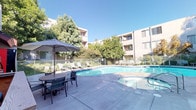 This outdoor amenity space centers around a large, freeform swimming pool with crystal-clear blue water, designed to invite relaxation and recreation. Adjoining the main pool is a dedicated spa, offering a separate space for hydrotherapy. The expansive concrete pool deck provides ample room for lounging, furnished with multiple chaise lounge chairs positioned to catch the sun, as well as a shaded dining area complete with an umbrella-covered table and chairs — ideal for outdoor gatherings. A vending machine is conveniently located near the seating area, adding a practical touch to the amenity space. The entire area is enclosed by a secure wrought iron fence with a gated entry, ensuring privacy and safety for users. Mature deciduous trees and tropical palms are planted throughout and along the perimeter, providing natural shade and a verdant backdrop against the multi-story stucco residential buildings that frame the courtyard. The open-sky setting allows for abundant natural light throughout the day, creating a bright and welcoming atmosphere that is well-suited for both active recreation and quiet outdoor relaxation.