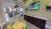 The Kids Cave is a thoughtfully designed children's play area that combines fun and functionality in a compact, single-floor layout. The room features bright, sage-green accent walls adorned with whimsical animal-themed artwork and a large wall-mounted flat-screen television, creating an engaging and entertaining environment for young guests. A child-sized round yellow table with vibrant lime-green frog-shaped chairs sits atop a colorful striped area rug, offering a dedicated space for play and activities. Built-in dark espresso cabinetry with a speckled granite countertop lines one wall, providing practical storage and surface space. The flooring throughout is a durable wood-look plank material that complements the overall modern aesthetic. Floor-to-ceiling glass partition walls with sleek metal framing define the space while maintaining an open, transparent feel, allowing clear sightlines into the adjacent fitness center and yoga studio. This connectivity ensures that parents can keep watch over children while utilizing nearby amenities. Recessed ceiling lighting provides a well-lit atmosphere, contributing to the room's clean, polished appearance. The Kids Cave strikes a balance between a safe, stimulating retreat for children and a seamlessly integrated component of a larger amenity suite.